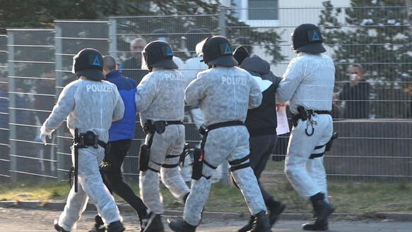Vier Polizeibeamte mit Helm und in weißen Ganzkörperschutzanzügen führen zwei Männer ab.