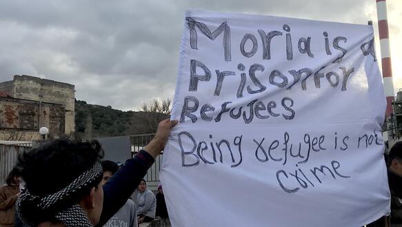 Personen blicken von der Kamera weg und halten ein weißes Banner hoch, auf dem steht: "Moria is a Prison for Refugees. Being refugee is not a crime."