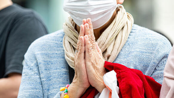 Eine ältere Frau mit Mundschutz faltet ihre Hände zum Gebet