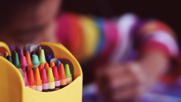 Box mit Stiften im Vordergrund. Im Hintergrund malt ein Kind mit buntem Pullover auf ein Papier.