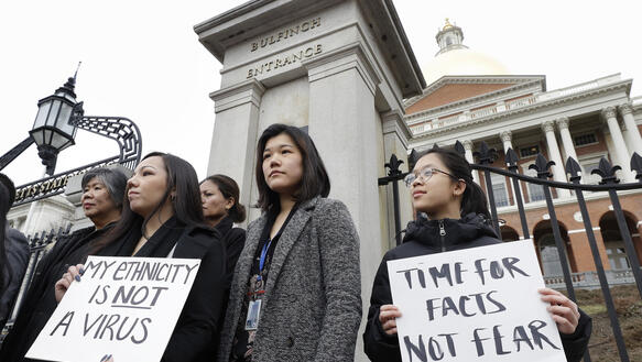 Mehrere Frauen stehen vor einem Gebäude und demonstrieren, sie halten Schilder mit der Aufschrift "Meine Ethnizität ist kein Virus" und "Zeit für Fakten, nicht für Angst"