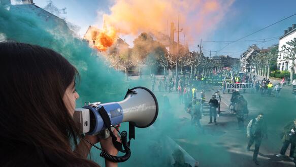 Im Vordergrund links eine Person mit Megaphon, im Hintergrund ein öffentlicher Platz mit Menschen die demonstrieren, blauer und orangener Rauch