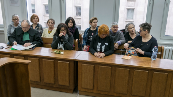 Szene aus einem Gerichtssaal, eine Gruppe von Frauen sitzen auf mehreren Bänken, daneben ein Mann in Robe
