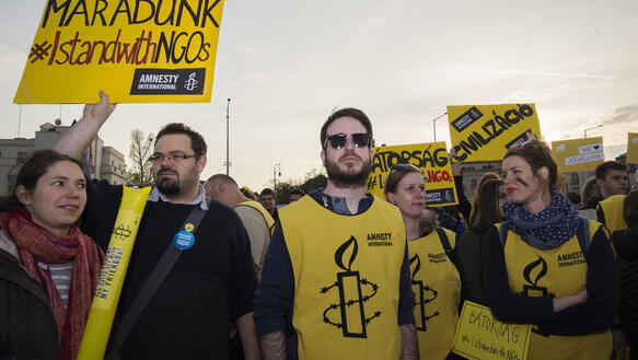 Personen in gelben Amnesty-Westen mit gelben Schildern, auf denen u.a. "#IstandwithNGOs" steht