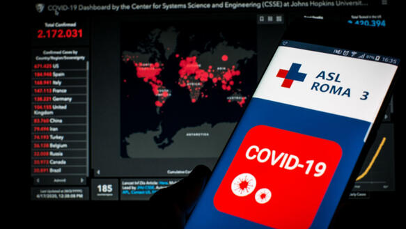 Eine Hand hält ein Smartphone auf dessen Display eine Covid-19-App angezeigt wird. Im Hintergrund der Illustration wird eine Weltkarte mit aktuellen Infektionszahlen angezeigt.
