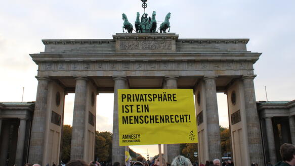 Eine Menschenmenge steht vor dem Brandenburger Tor. Eine Person hält ein Schild mit Amnesty-Logo hoch. Auf dem Schild steht: "Privatsphäre ist ein Menschenrecht!".