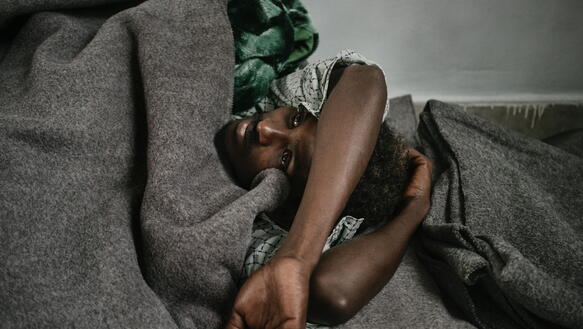 Ein Schwarzer Mann liegt zugedeckt mit einer grauen Wolldecke auf dem Boden und schaut in sich versunken in die Höhe