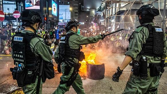 Drei uniformierte Polizisten mit Schlagstöcken sind auf einer Straße positioniert. Im Hintergrund befindet sich ein Feuer, Protestierende und moderne Häuserfassaden.