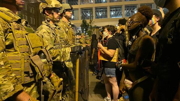Auf einer Straße steht eine Reihe uniformierter Soldaten mit Schutzschildern einer Reihe von Demonstrant_innen gegenüber.