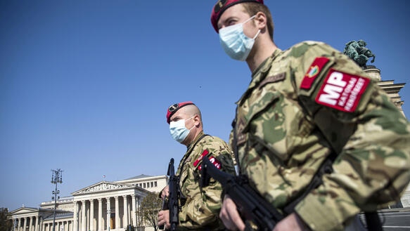 Zwei Männer mit Mundnasenschutzmasken in Militäruniform halten Maschinenpistolen in ihren Händen und stehen vor einem Gebäude.