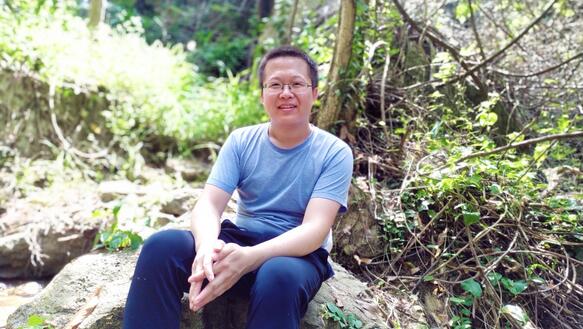 Ein junger Mann sitzt auf einem Stein im Wald und lächelt in die Kamera.