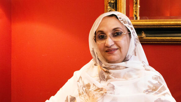 Eine mittelalte Frau mit Brille und Kopftuch, es ist die Menschrenrechtsverteidigerin Aminatou Haidar, sitzt vor einer roten Wand an der ein goldgerahmter Spiegel hängt.