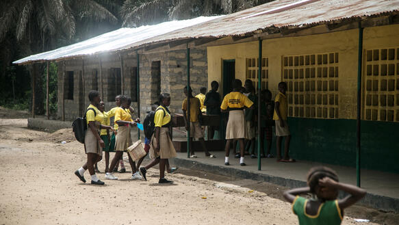 Mehrere Schülerinnen stehen vor einfachen Schulräumen, sie tragen gelbe T-Shirts, die Schulgebäude haben ein Dach aus Wellblech, dahinter zu sehen sind Palmen