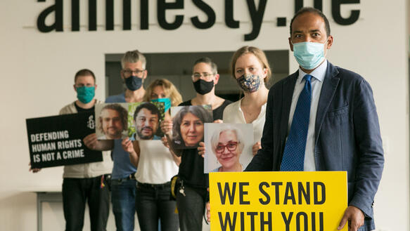 Eine Menschengruppe mit Protestschildern "We stand with you" und Porträts in den Händen, sowie Mundschutzmasken steht in einem Raum und guckt in die Kamera. Im Hintergrund an der Wand steht: amnesty.de