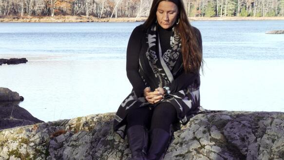 Sherri Mitchell sitzt auf einem Stein am Ufer eines Sees und blickt auf ihre in ihrem Schoß gefalteten Hände.
