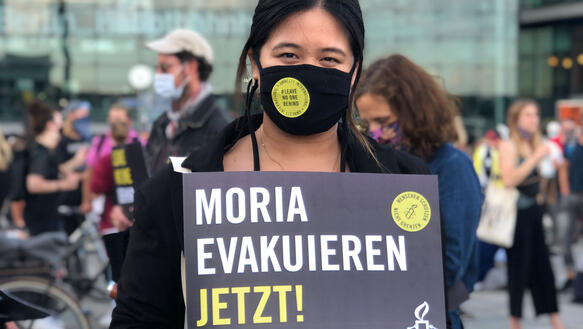 Eine junge Frau mit Mundschutz steht in einer Menschenmenge und hält ein Schild in der Hand, auf dem steht: "Moria evakuieren. Jetzt!". Auf dem Aufkleber auf ihrem Mundschutz steht: "Amnesty International. Leave no one behind".