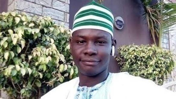 Porträtfoto von Yahaya Sheriff-Aminu, aufgenommen vor einer Hecke vor einen Gebäude. Er trägt eine traditionelle Kopfbedeckung und ein traditionelles Gewand und lächelt in die Kamera.