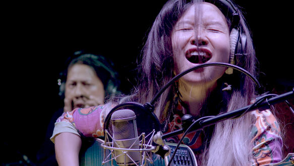 Eine junge Frau trägt Kopfhörer und singt mit geschlossenen Augen, während sie Gitarre spielt.