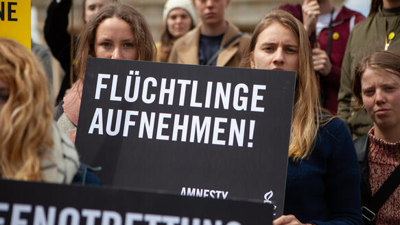 Zwei junge Frauen blicken in die Kamera und halten ein Schild in der Hand, auf dem steht: "Flüchtlinge aufnehmen". Hinter ihnen stehen weitere Personen, von denen einige ebenfalls Schilder hochhalten.