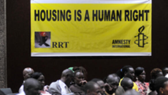 Menschen sitzen am Boden vor einem gelben Plakat, auf dem in schwarzen Lettern "Housing is a human right" steht.