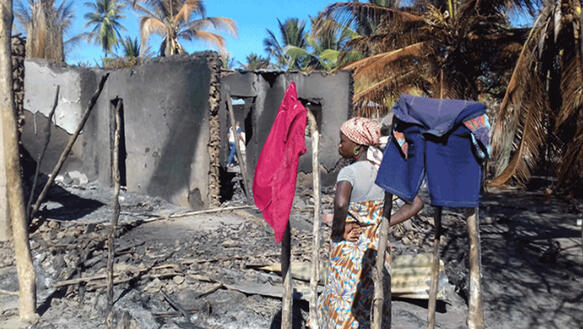 Eine Frau steht inmitten von Trümmern eines abgebrannten Hauses, Palmbäume daneben sind ebenfalls verkohlt