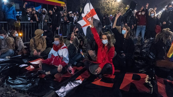 Mehrere Frauen sitzen mit Mund-Nasen-Masken auf einem großen Banner, sie heben die Hand zum Victory-Zeichen, es ist bereits Nacht.