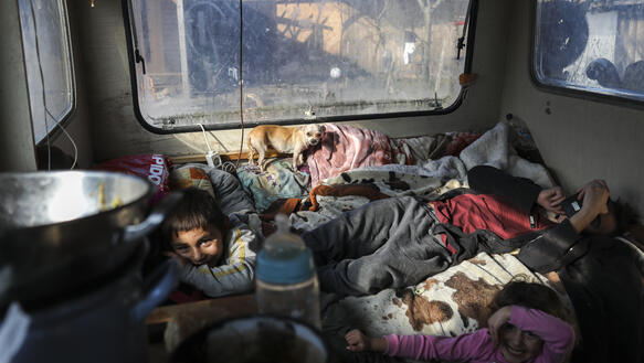 Innenansicht eines älteren Wohnwagens, auf dem Bett sieht man Decken, mehrere Kinder, die in die Kamera schauen, ein Hund und einen Mann mit Hand