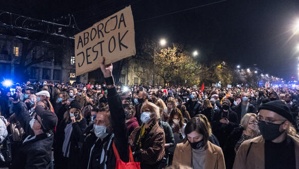 Viele Männer und Frauen, die Mundnasenschutz tragen, stehen bei Dunkelheit auf einer Straße und demonstrieren; ein Mann hält ein Pappschild hoch, auf dem in polnisch geschrieben steht: "Abtreibung ist ok".