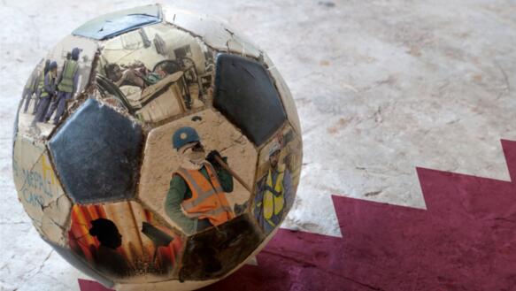 Ein Fußball liegt auf der Flagge Katars. Auf dem Fußball sind Fotos abgedruckt, die Bauarbeiter und Hausangestellte bei ihrer Arbeit oder in ihren Unterkünften zeigen.
