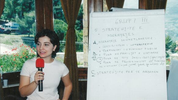 Eine junge Frau mit einem roten Mikrofon, daneben eine Informationstafel