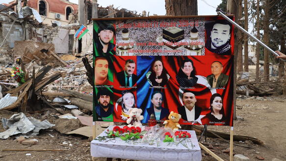 Eine Tafel mit zehn Porträtbildern steht vor zerstörten Gebäuden. Vor der Tafel steht ein Tisch, auf dem Blumen und Teddybären liegen.274128