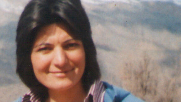 Eine Frau mit schulterlangen schwarzen Haaren und blauer Jacke lächelt in die Kamera. Die Sonne scheint und im Hintergrund befinden sich ein Tal und Berge.