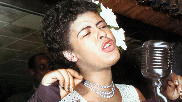 Eine schwarze Frau mit Perlenkette singt in ein Mikrofon und neigt dabei den Kopf etwas zur Seite.