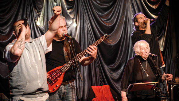 Drei mittelalte Männer und eine ältere Dame stehen auf einer Bühne vor einem Vorhang und musizieren; einer von ihnen spielt Bass, alle von ihnen singen in Mikrofone.