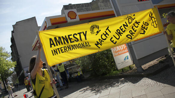 Eine Frau und ein Mann in Amnesty-Kleidung halten vor einer Tankstelle mit dem Shell-Logo ein Transparent hoch, auf dem steht: "Macht euren Dreck weg!".