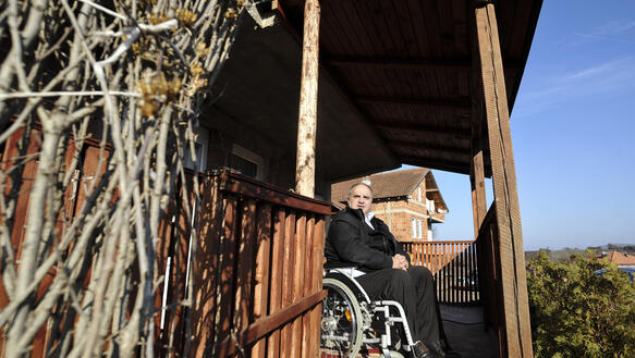 Ein Mann sitzt in einem Rollstuhl auf der Holzveranda seines Hauses und trägt eine dicke Winterjacke; im Hintergrund steht ein unverputztes Haus.