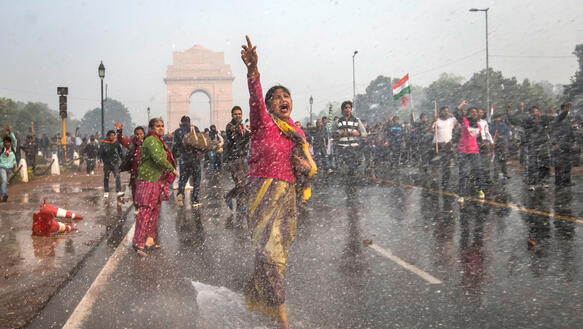 Eine Frau läuft vorm India Gate in Neu-Delhi die Straße entlang. Sie hat ihre rechte Hand erhoben und ruft etwas. Dabei treffen sie Wassertropfen, potenziell von einem Wasserwerfer. Hinter ihr befinden sich weitere Protestierende.