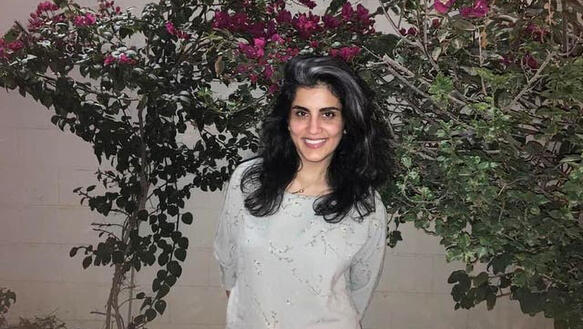 Loujain Al-Hathloul lächelt in die Kamera, sie trägt ihr teilweise leicht ergrautes Haar offen. Sie steht vor einer Mauer, die mit blühenden Blumensträuchern bedeckt ist.