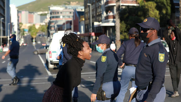 Eine Frau schreit auf einer Straße eine Gruppe Polizisten an, die Schirmmützen und Mund-Nase-Schutz tragen.