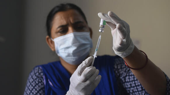 Das Foto zeigt eine indische Frau mit Atemschutzmaske und Handschuhen, die eine Spritze mit Impfstoff füllt.