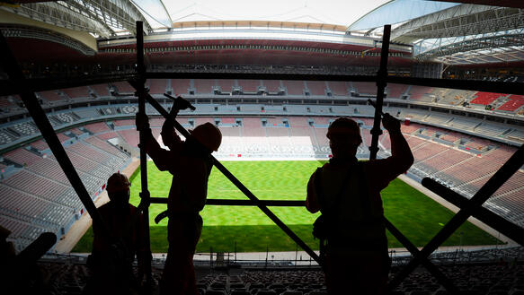 Das Bild zeigt das Innere eines Fußballstadion, im Vordergrund die Silhouetten mehrere Bauarbeiter, die ein Baugerüst abbauen