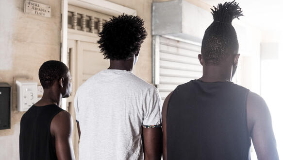 Das Bild zeigt drei Jugendliche in einem Hausflur, sie blicken in Richtung eines Fensters, man sieht ihre Gesichter nicht.