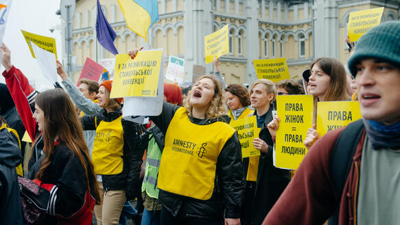 Verschiedene Menschen, darunter Amnesty-Unterstützende mit gelben Westen und dem Amnesty-Logo, demonstrieren.