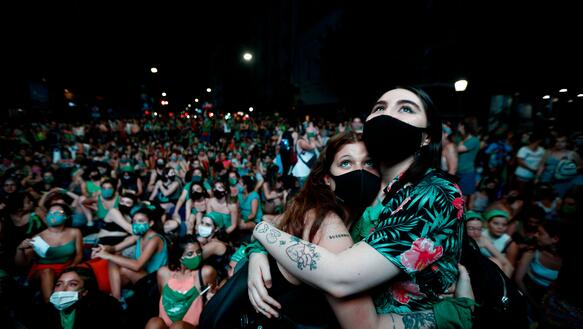 Zwei Frauen umarmen sich mit Masken im Vordergrund. Hinter ihnen befinden sich viele weitere Demonstrantinnen.