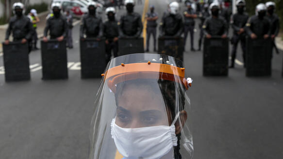 Das Foto zeigt eine Frau mit Faceschild und Gesichtsmaske auf einer Straße. Im Hintergrund stehen eine Reihe bewaffneter Polizisten.