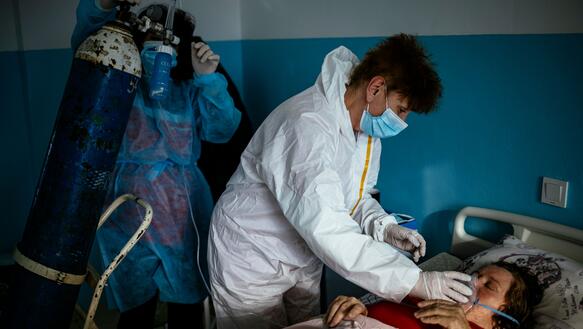 Das Bild zeigt eine Frau in einem Krankenbett. Zwei Pflegerinnen in medizinischen Schutzanzügen setzen ihr eine Schutzmaske auf.