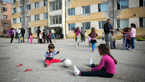 Kinder sitzen auf dem Boden und spielen Ball, im Hintergrund Menschen und ein Haus in schlechtem Zustand