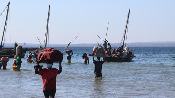 Das Bild zeigt einen Strand und das Meer, mehrere Personen gehen mit Gepäckstücken auf dem Kopf von kleinen Segelbooten in Richtung Strand