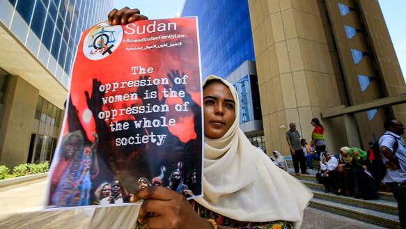 Das Bild zeigt eine Frau mit Kopftuch, die ein Plakat in der Hand hält mit der Aufschrift "The oppression of women is the oppression of the whole society"