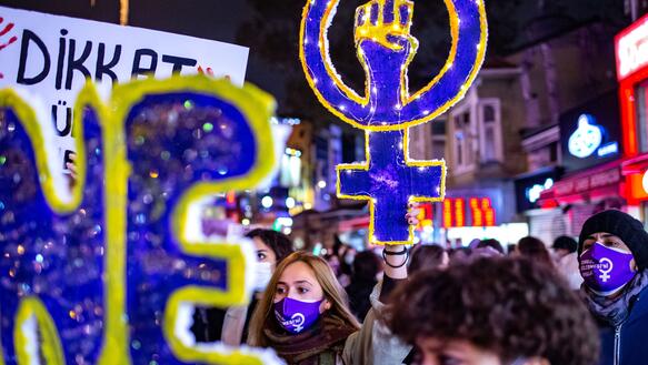 Eine junge Frau mit Mundschutz hält inmitten einer demonstrierenden Menschenmenge ein gebasteltes Plakat in Form des Weiblich-Symbols hoch. Der Kreis des Symbols enthält eine zur Faust geballte Hand.